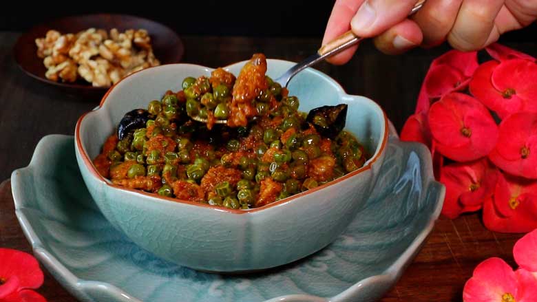 A hand is seen holding a spoon and picking up Malvani green peas and walnut curry from a blue celadon bowl lying on a blue celadon scalloped lotus platter with california walnuts and red flowers in the background - Malvani Green Peas and Walnut Curry Recipe